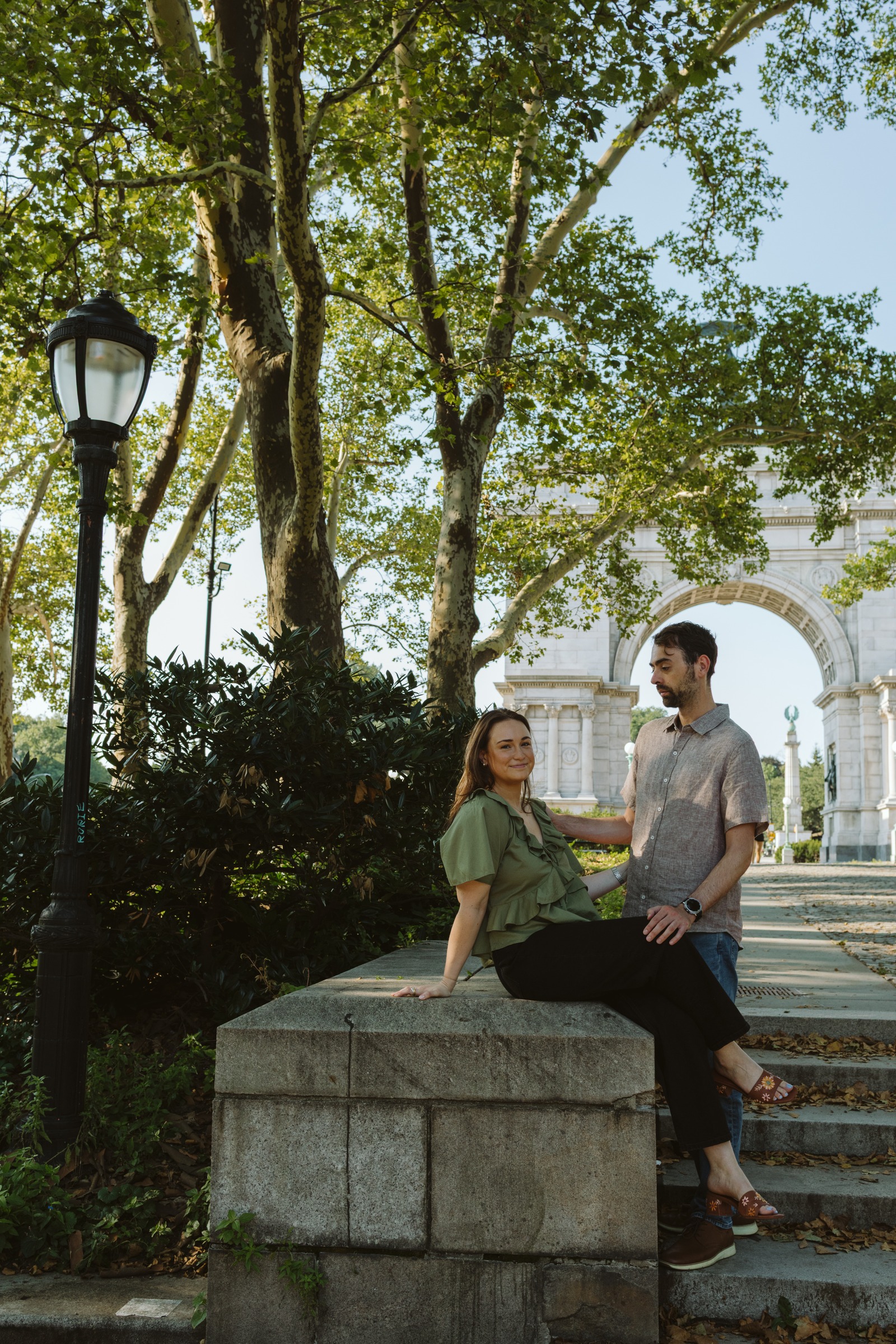 Monika sitting on a stone ledge in Grand Army Plaza with Haggai standing beside her, the memorial arch behind them.