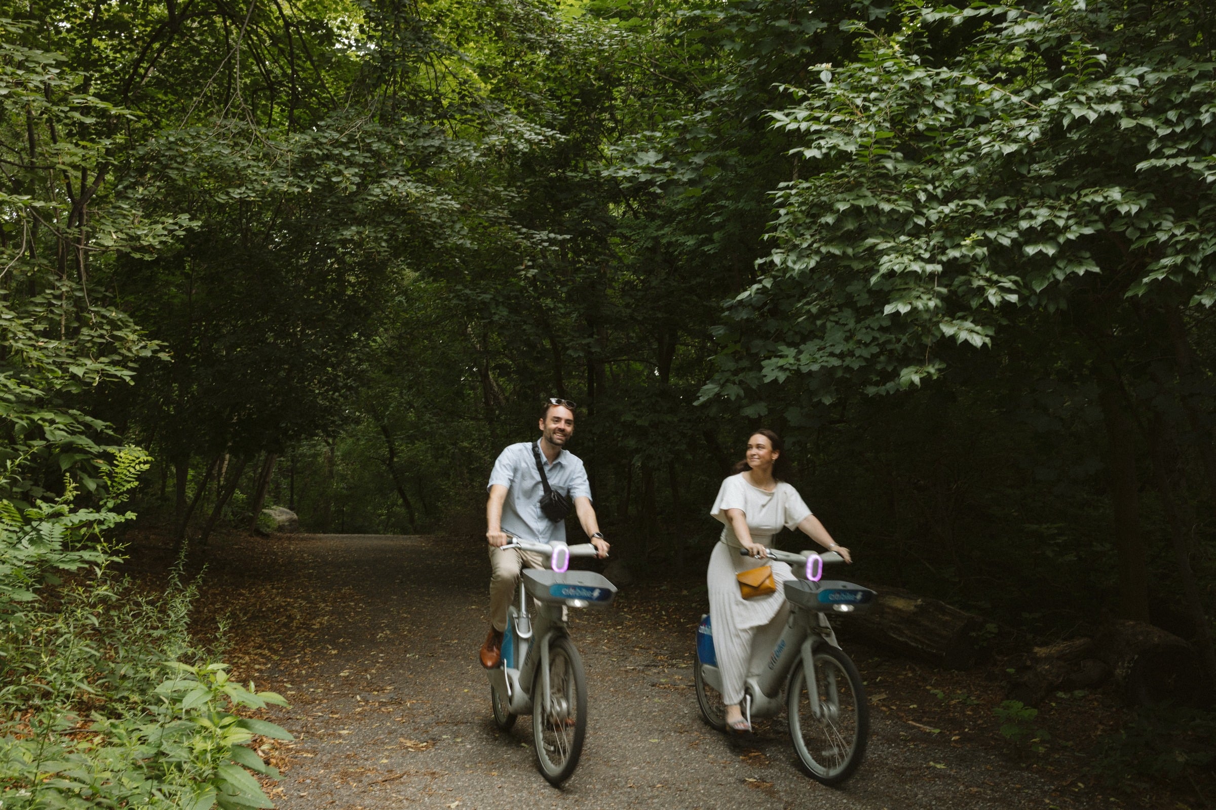 Walking their bikes side by side down a shaded path through the park.